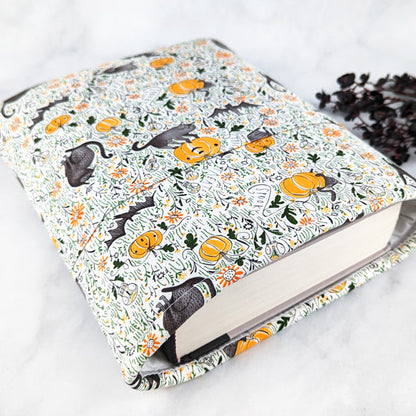 Fabric book sleeve with a cream background and a Halloween-themed pattern of black cats, bats, pumpkins, mushrooms, and orange flowers scattered among green vines.