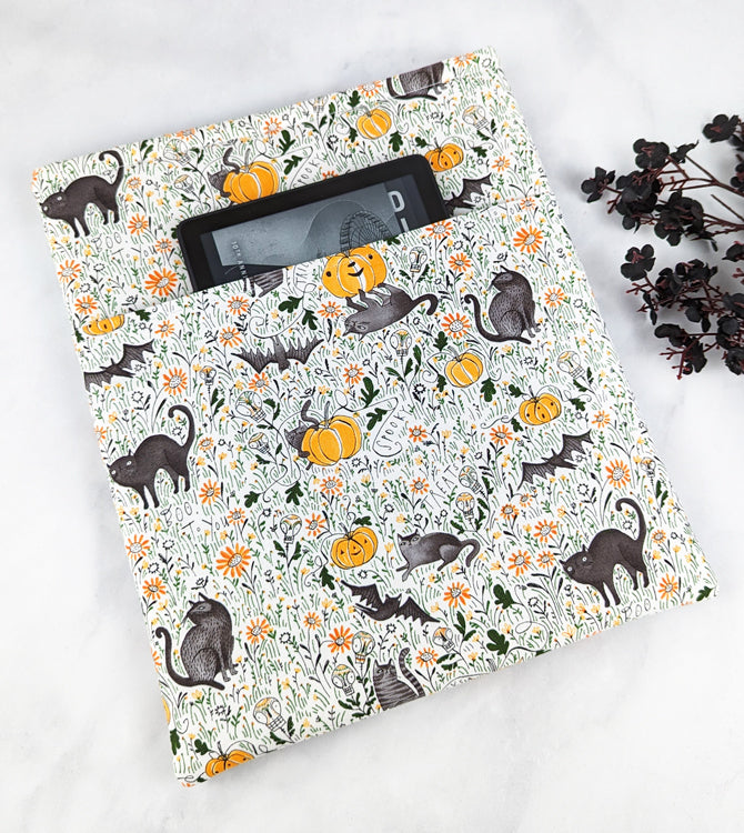 Fabric book sleeve with a cream background and a Halloween-themed pattern of black cats, bats, pumpkins, mushrooms, and orange flowers scattered among green vines.