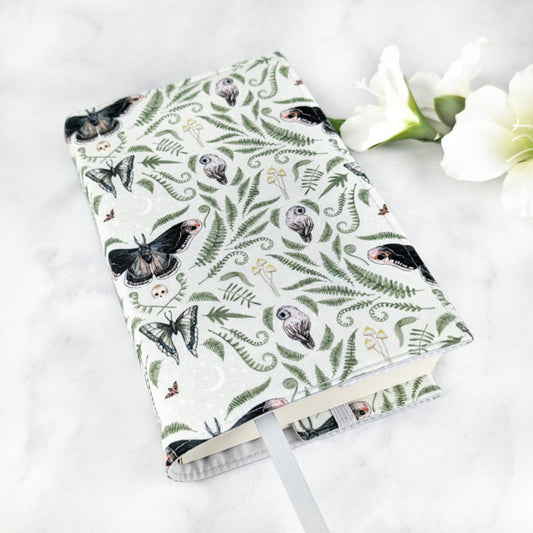Fabric book cover with a mystical forest design featuring detailed illustrations of owls, moths, butterflies, ferns, mushrooms, and crescent moons on a soft sage green background, finished with a pale gray ribbon bookmark.