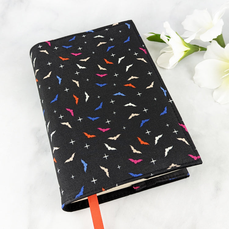 medium adjustable book cover, fabric features mini bat shapes in shade of orange, blue, pink and white, on a black background.