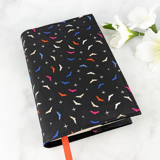 medium adjustable book cover, fabric features mini bat shapes in shade of orange, blue, pink and white, on a black background.