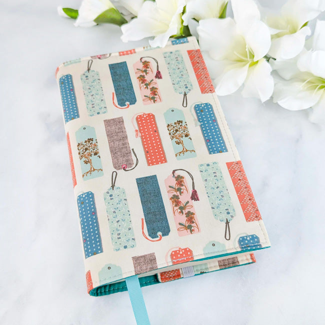 Fabric book cover featuring a pattern of colorful vintage-style bookmarks on a cream background, fitted on a book with a light blue ribbon bookmark, displayed beside white flowers.