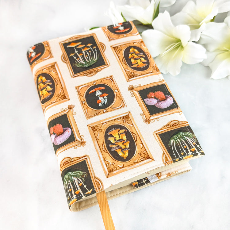 Cream book cover featuring framed mushroom illustrations in golden ornate borders, creating a whimsical naturalist gallery design.