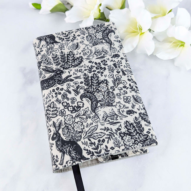 Fabric book cover with a black and white woodland pattern featuring rabbits, squirrels, and floral elements, fitted on a book with a black ribbon bookmark, displayed beside white flowers.