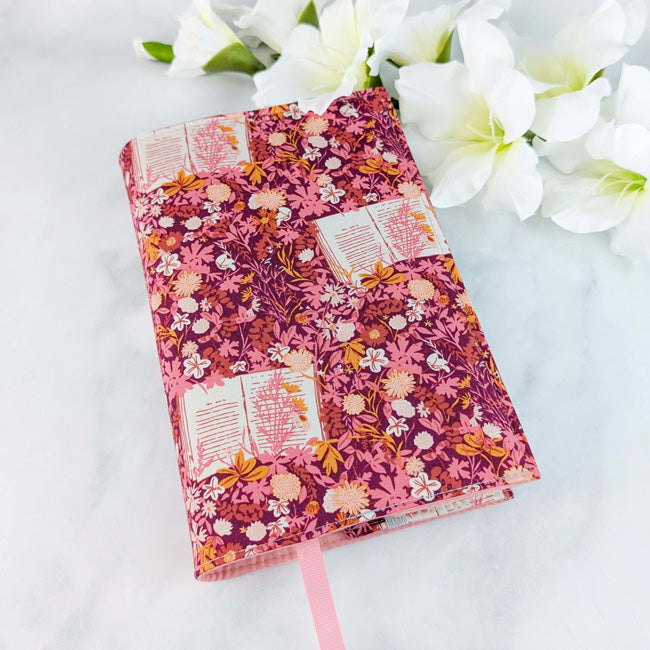 Fabric book cover with a rich pink and burgundy floral pattern featuring open books and delicate flowers, fitted on a book with a pink ribbon bookmark, displayed beside white flowers.