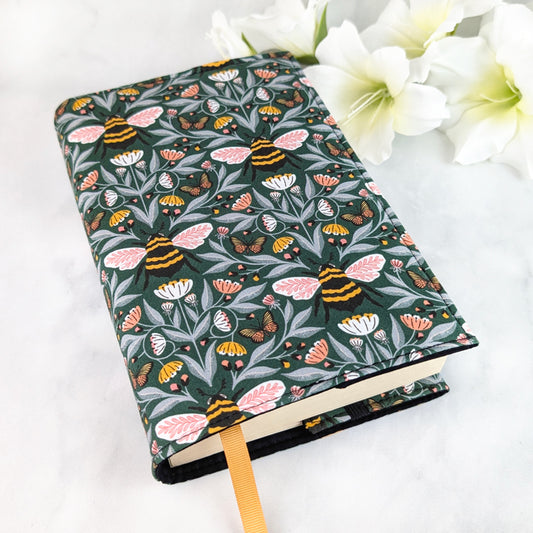 Dark green book cover with a detailed bee and butterfly pattern surrounded by pink, white, and yellow wildflowers on leafy vines.