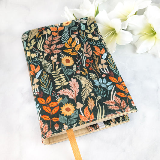 Dark green book cover featuring an intricate woodland pattern with autumn leaves, sunflowers, mushrooms, and ferns in warm orange, gold, and muted blue tones.