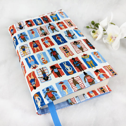 adjustable fabric book cover, fabric pattern features multiple illustrations of women of various races laying on colorful beach towels.