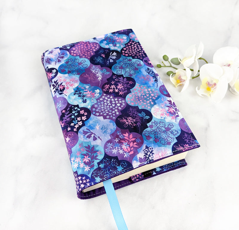 Fabric book cover with a purple, blue, and pink floral medallion pattern, fitted on a book with a light blue ribbon bookmark, displayed beside white flowers.