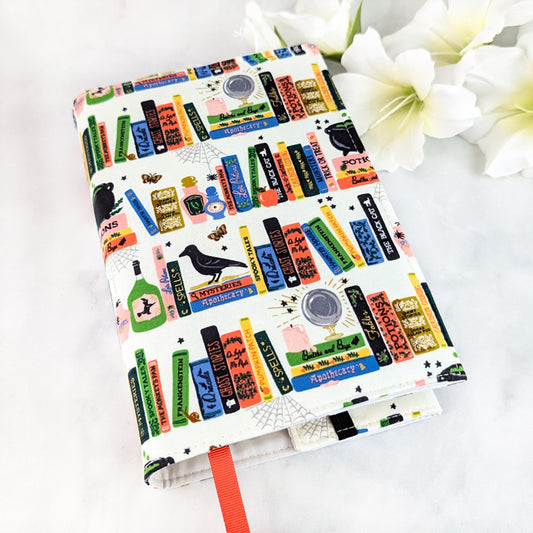 Off-white book cover decorated with colorful illustrated books, potion bottles, ravens, and spell labels in a playful witchy library design.