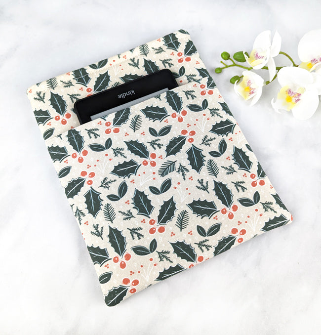Cream-colored fabric book sleeve with a festive holly and berry print in dark green and red, shown holding a hardcover book on a white marble background with white orchids nearby.