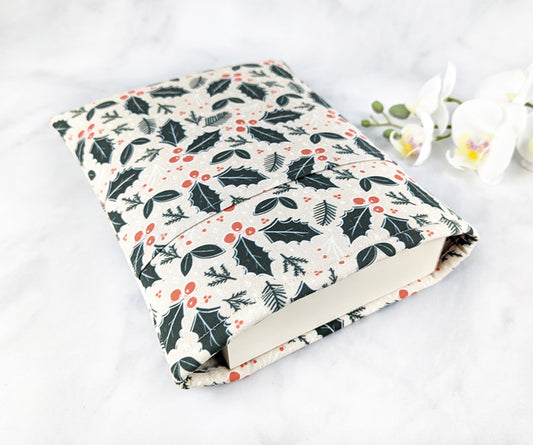 Cream-colored fabric book sleeve with a festive holly and berry print in dark green and red, shown holding a hardcover book on a white marble background with white orchids nearby.