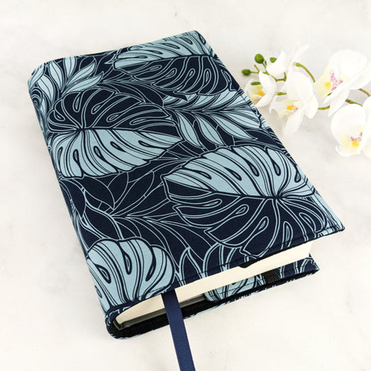 Fabric book cover with a bold tropical design featuring large monstera leaves in light blue outlines on a deep navy background, finished with a matching navy ribbon bookmark.