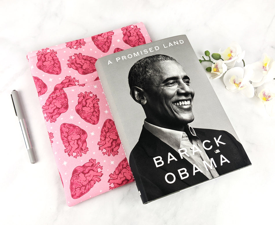 A pink fabric book sleeve featuring a repeating pattern of red anatomical hearts and white stars. Hardcover version of "A Promised Land" by Barack Obama laying next to it for scale.