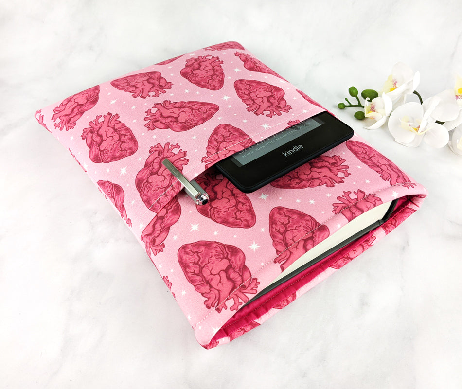 A pink fabric book sleeve featuring a repeating pattern of red anatomical hearts and white stars. Pen and Kindle sleeve inside the side pocket.