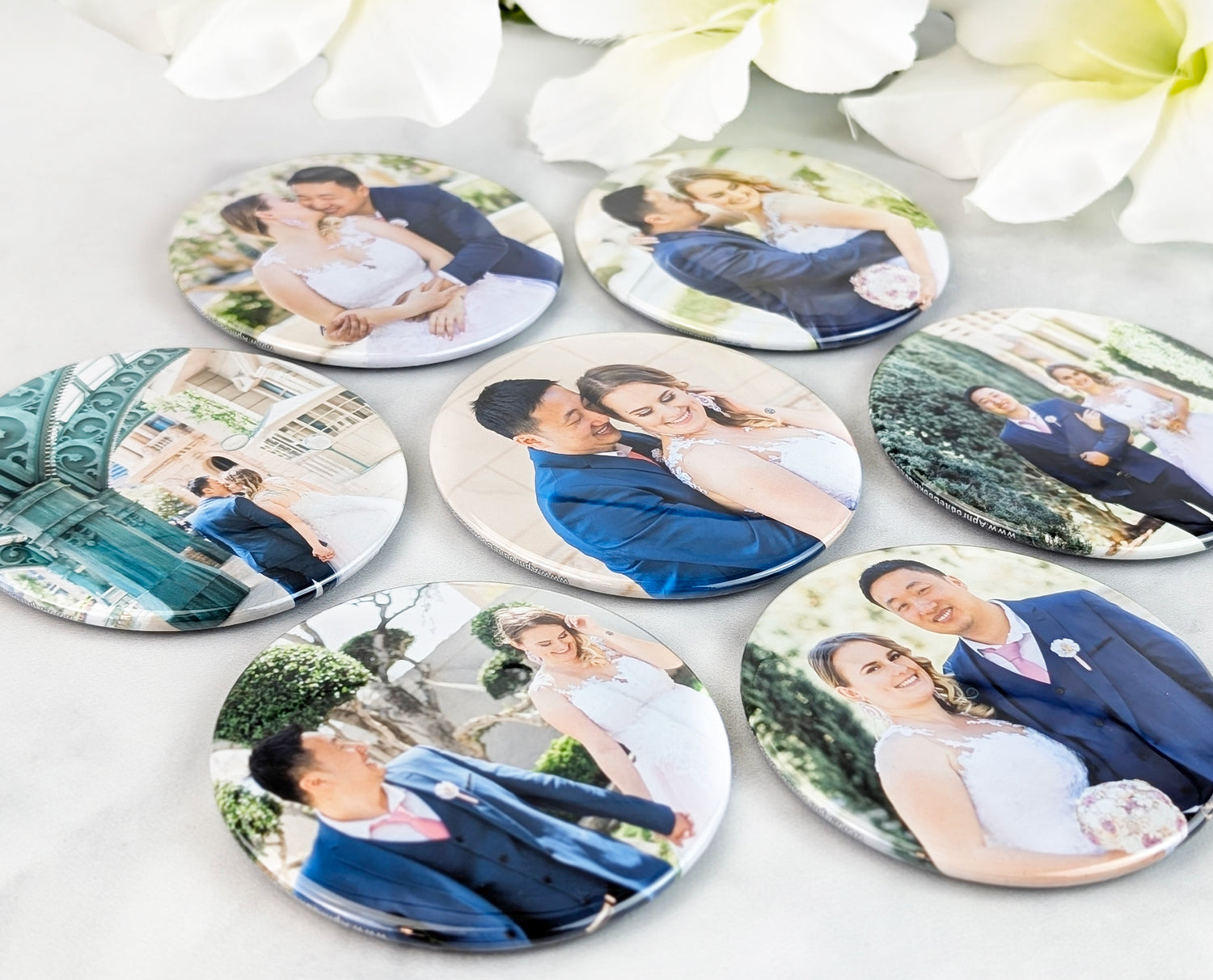 A set of round photo magnets featuring vibrant wedding portraits of a couple in various romantic poses. The magnets are arranged on a soft, light surface and accented with white flowers, showcasing the glossy finish and vivid, full-color printing.