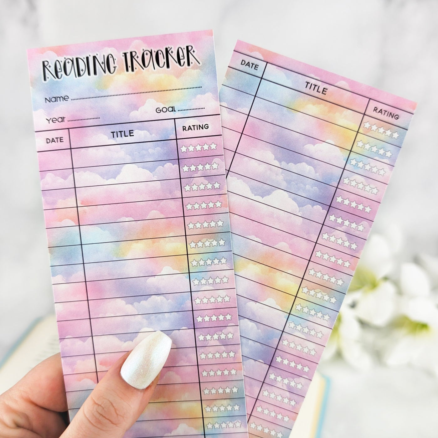 hand holding two bookmark reading trackers, one showing front and one the back. bookmark has a pastel cloud background and the table to track and rate books read.