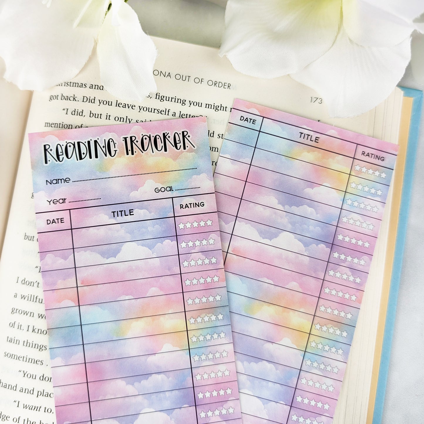 two bookmark reading trackers laying on a page of an open book, one showing front and one the back. bookmark has a pastel cloud background and the table to track and rate books read.