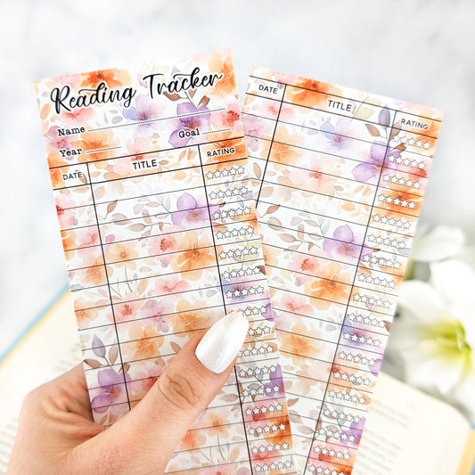 hand holding two bookmark reading trackers, one showing front and one the back. bookmark has a pastel floral background in orange, pink and purple color, and the table to track and rate books read.