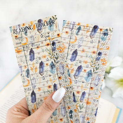 hand holding two bookmark reading trackers, one showing front and one the back. bookmark has a witchy background with moons, crystals and greenery, and the table to track and rate books read.
