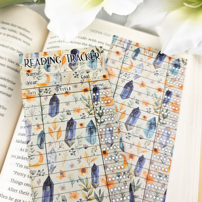 two bookmark reading trackers laying on a page of an open book, one showing front and one the back. bookmark has a witchy background with moons, crystals and greenery, and the table to track and rate books read.