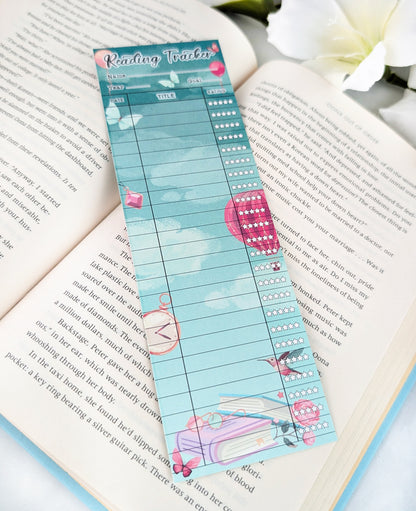 Whimsical teal reading tracker bookmark with balloons, butterflies, and books design, placed on an open book.
