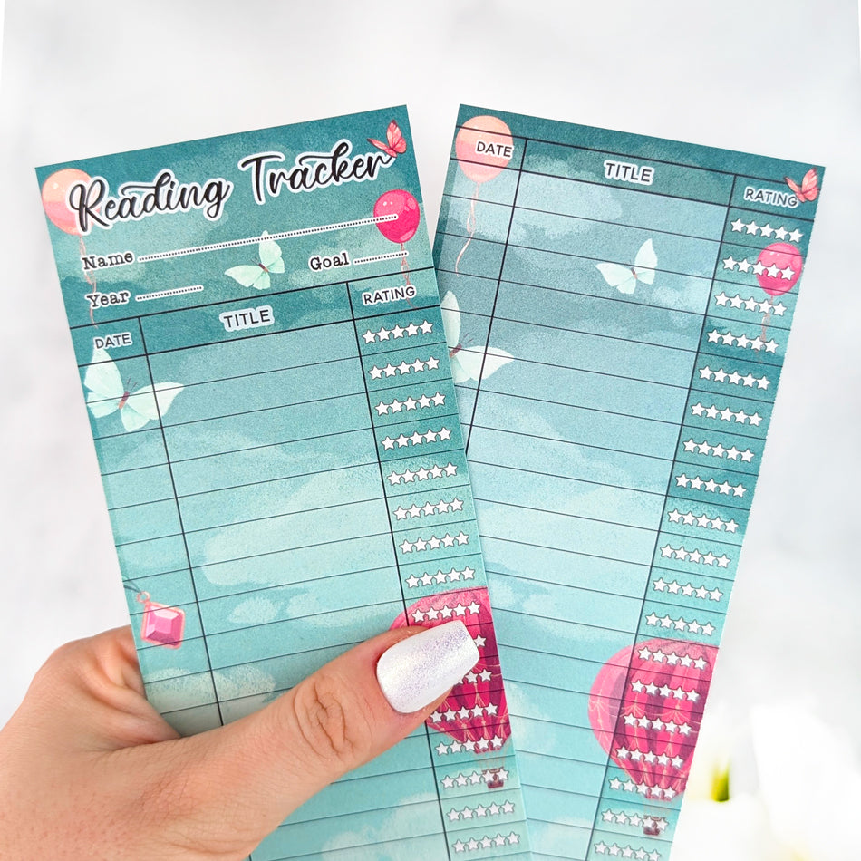 Two teal fantasy-themed reading tracker bookmarks with balloon and butterfly accents, held in a hand with silver nail polish.