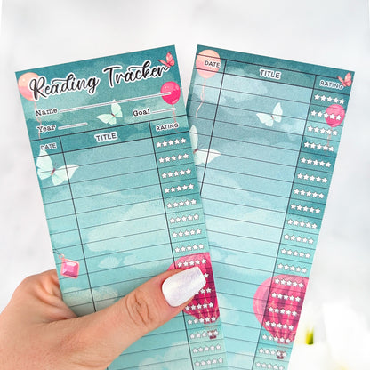 Two teal fantasy-themed reading tracker bookmarks with balloon and butterfly accents, held in a hand with silver nail polish.
