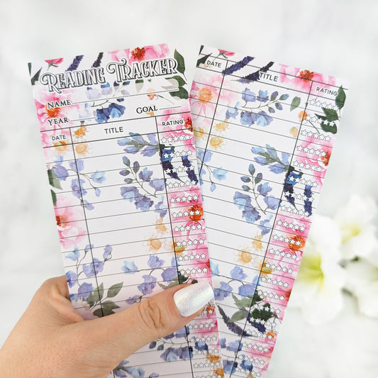 Two floral reading tracker bookmarks with vibrant bouquet designs, held in a hand with silver nail polish.