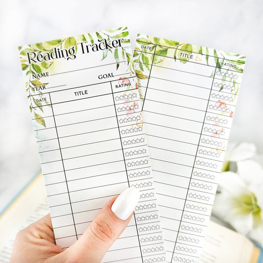 hand holding two bookmark reading trackers, one showing front and one the back. bookmark has a white background and floral greenery adorning edges of the bookmark, and the table to track and rate books read.