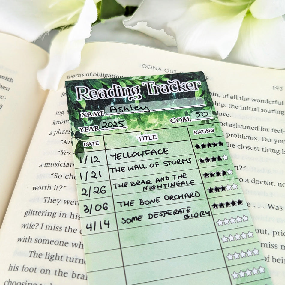 Green forest-inspired reading tracker bookmark with handwritten book titles and star ratings, resting on an open book.