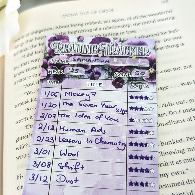 Close-up of a purple floral reading tracker bookmark filled out with handwritten book titles, dates, and star ratings, placed on an open book.