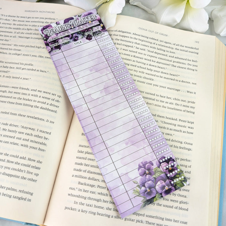 Purple floral reading tracker bookmark with star rating columns placed on an open book, decorated with pansy flowers at the top and bottom.