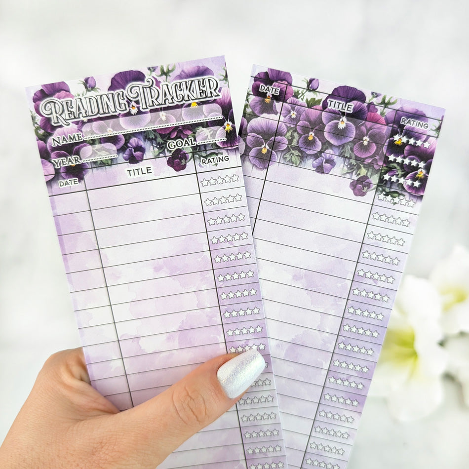 Two purple floral reading tracker bookmarks with lined sections for date, title, and star ratings, held in a hand with white pearly nail polish.