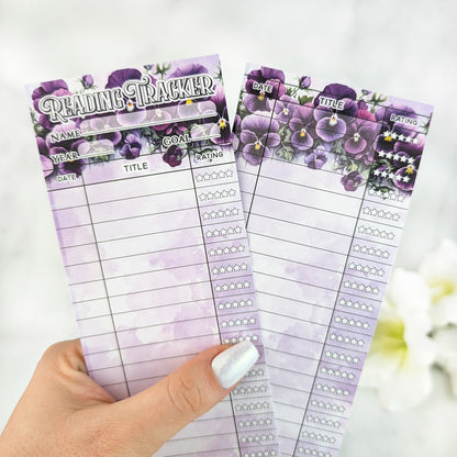Two purple floral reading tracker bookmarks with lined sections for date, title, and star ratings, held in a hand with white pearly nail polish.