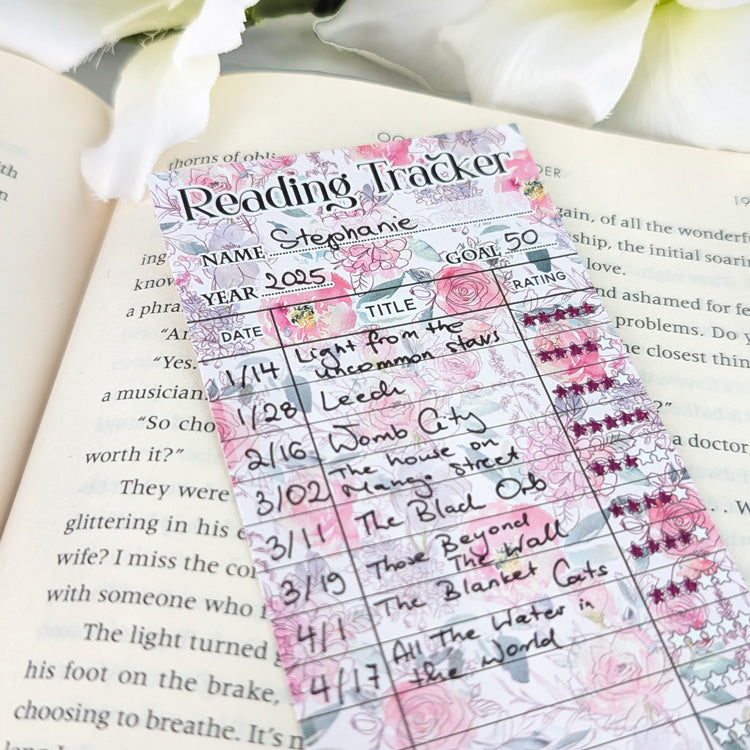 Close-up of a pastel floral reading tracker bookmark filled with handwritten book titles and star ratings, resting on an open book.