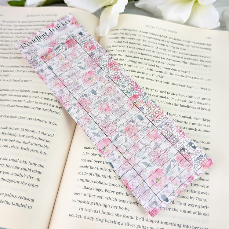 Pastel floral reading tracker bookmark with blank rows for dates, titles, and ratings, placed on an open book with white flowers nearby.