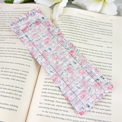 Pastel floral reading tracker bookmark with blank rows for dates, titles, and ratings, placed on an open book with white flowers nearby.