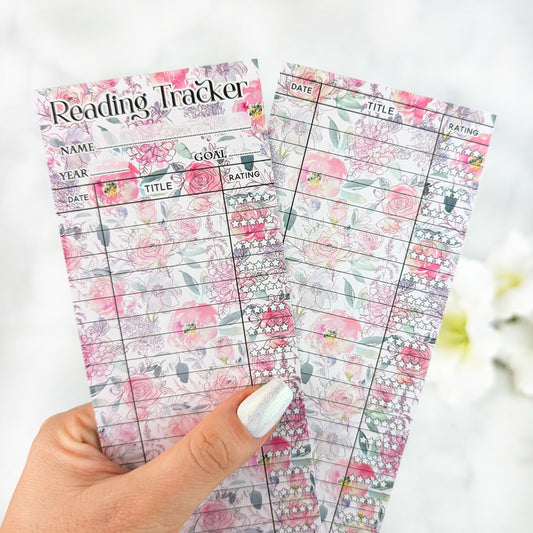Two pastel floral reading tracker bookmarks with pink rose designs, held in a hand with silver nail polish.