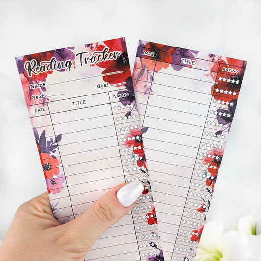 Two floral reading tracker bookmarks with bold red and purple flowers, held in a hand with silver nail polish.