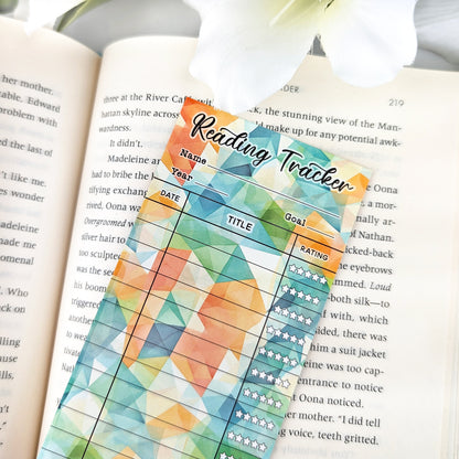 single bookmark reading tracker laying on an open book. bookmark has a geometric background in green, orange, yellow colors, and the table to track and rate books read.