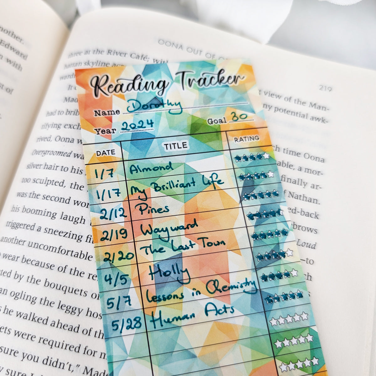 bookmark reading tracker laying on a page of an open book. bookmark has a geometric background in green, orange, yellow colors, and the table to track and rate books read. some of the titles are filled out.