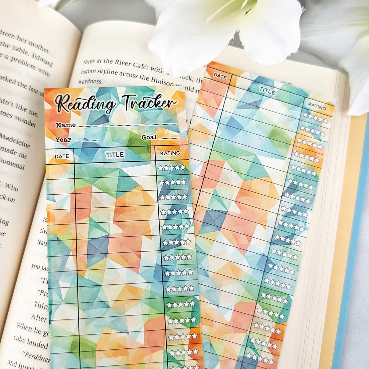 two bookmark reading trackers laying on a page of an open book, one showing front and one the back. bookmark has a geometric background in green, orange, yellow colors, and the table to track and rate books read.