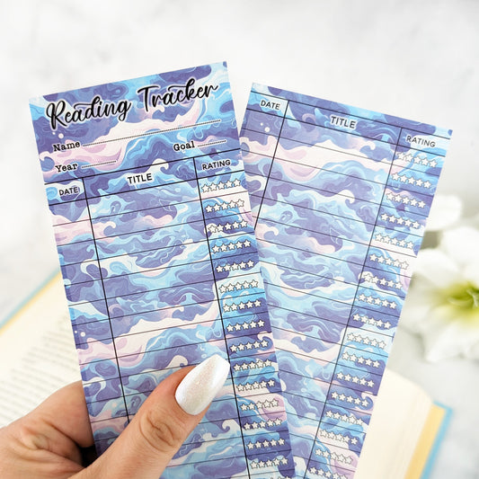 Two wave-pattern reading tracker bookmarks in shades of blue, purple, and pink, held in a hand with silver nail polish.