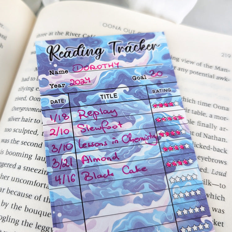 Blue and purple wave-pattern reading tracker bookmark filled with handwritten book titles and star ratings, resting on an open book.