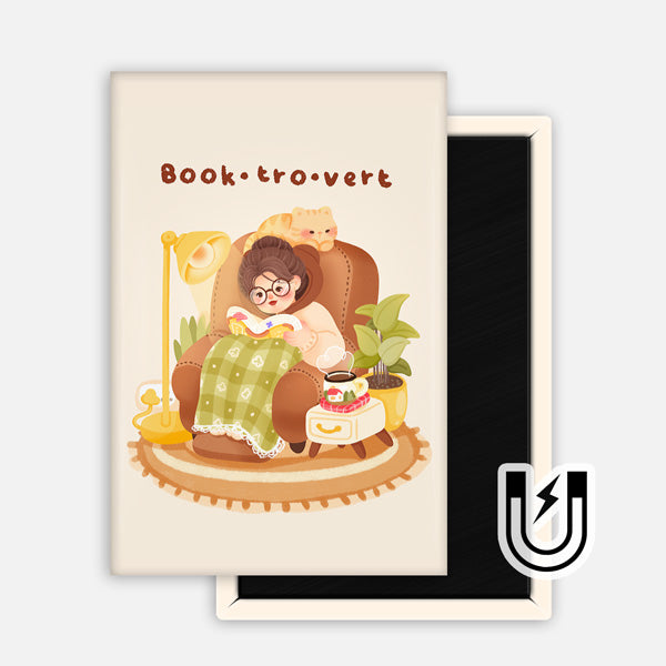 Cream-colored rectangular magnet featuring a cozy reader in glasses curled up in an armchair with a blanket, tea, and cat. Text reads “Book·tro·vert.”