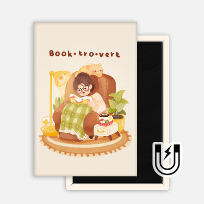 Cream-colored rectangular magnet featuring a cozy reader in glasses curled up in an armchair with a blanket, tea, and cat. Text reads “Book·tro·vert.”