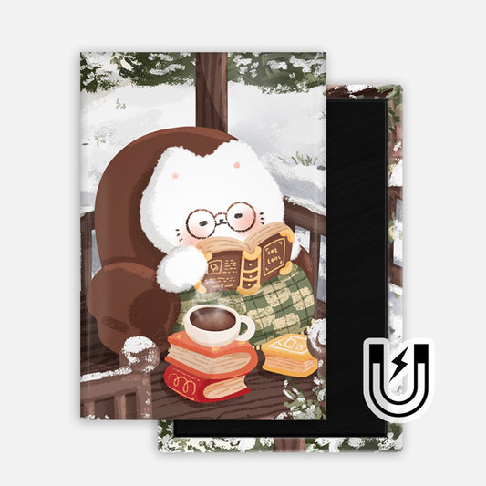 Rectangular magnet featuring a fluffy white cat wearing glasses, wrapped in a green blanket, reading a book on a snowy porch with a mug of cocoa and a stack of books beside it.