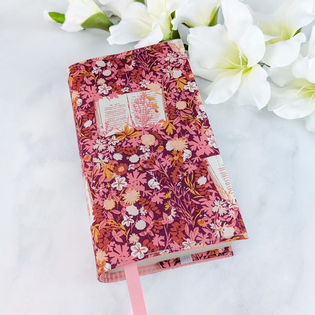 Fabric book cover with a rich pink and burgundy floral pattern featuring open books and delicate flowers, fitted on a book with a pink ribbon bookmark, displayed beside white flowers.