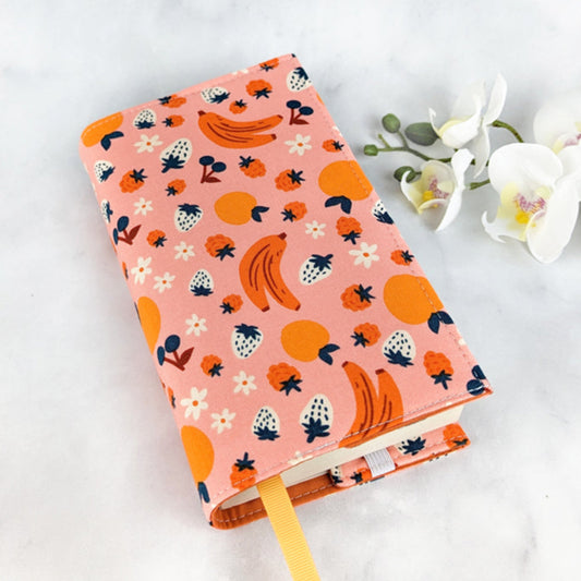 small adjustable book cover, fabric showcases oranges, bananas, strawberries, blueberries and flowers on a light pink background.